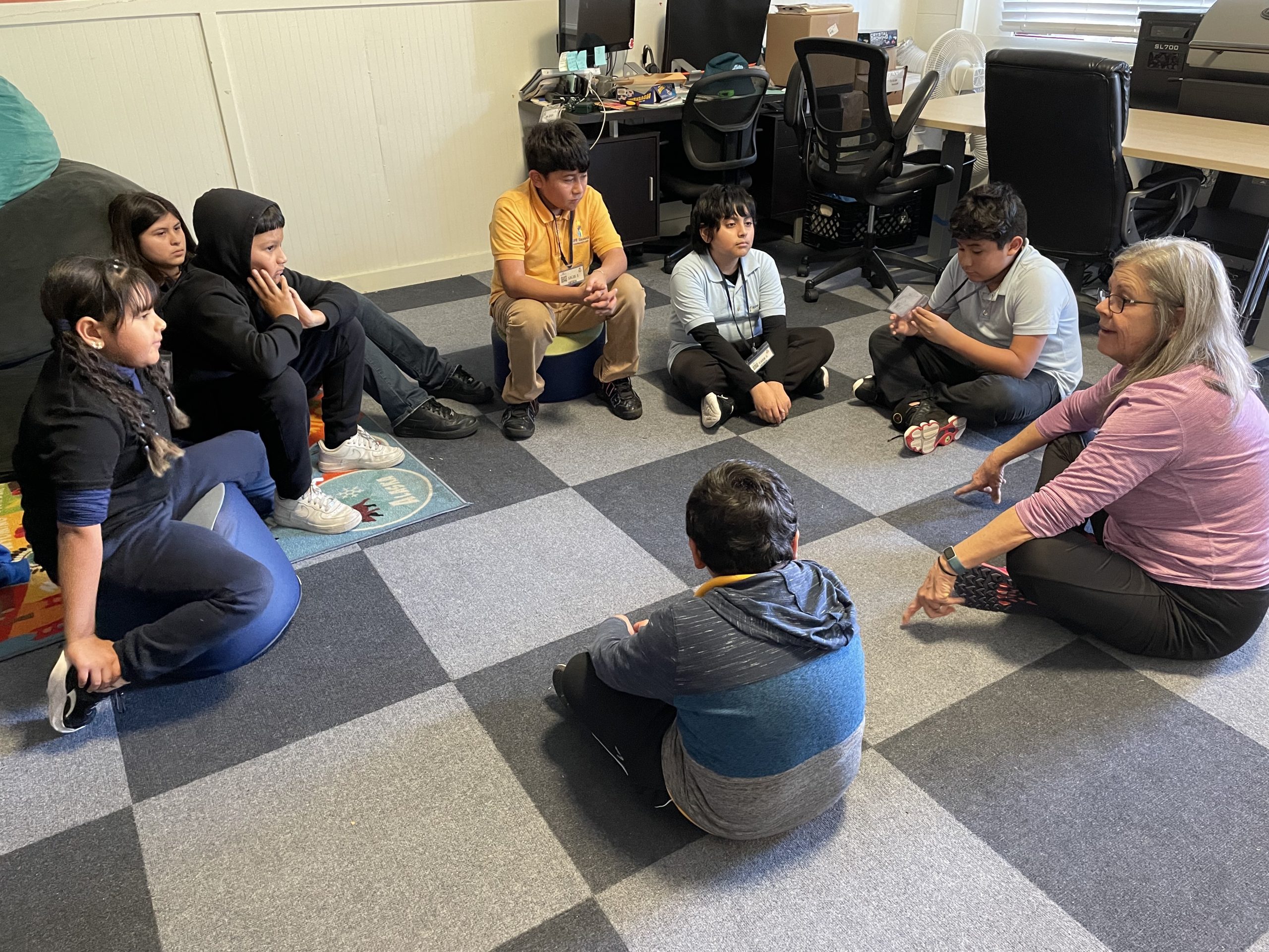 adult female volunteer sits cross-legged on the floor in a circle with 7 elementary school-aged children